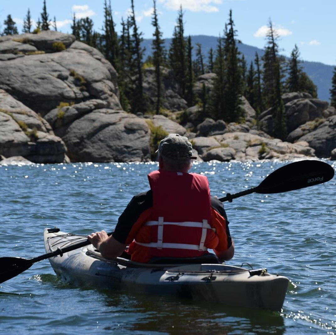 Person kayaking on a mountain lake surrounded by rocks and trees under a clear blue sky.